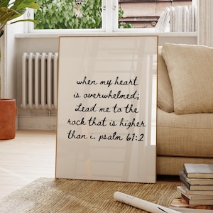 Peut inclure: Une affiche encadrée avec le texte "when my heart is overwhelmed; Lead me to the rock that is higher than i. psalm 61:2" en script noir. L'affiche est appuyée contre un canapé beige dans une pièce lumineuse avec une grande fenêtre et une plante en pot.