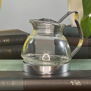 Puede incluir: Una cafetera de vidrio transparente con asa y base plateadas. La cafetera tiene un pequeño pico y una tapa con un pequeño pomo. La cafetera está sobre una pila de libros.