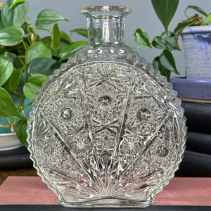 Puede incluir: Un jarrón de vidrio transparente con una base redonda y festoneada y un patrón geométrico texturizado. El jarrón tiene un cuello corto y una abertura ancha.