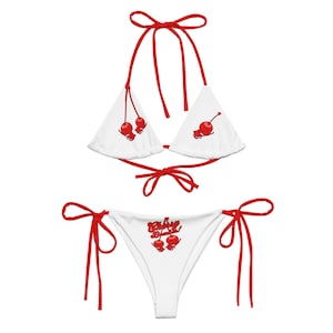 Könnte beinhalten: Weißes Bikini-Set mit roten Akzenten. Das Oberteil ist im Dreiecksstil mit roten Bindebändern, und das Unterteil ist ein klassischer Schnitt. Beide Teile zeigen eine rote Kirschgrafik. Auf dem Unterteil steht der Text "Cherry Bomb!" in Rot.