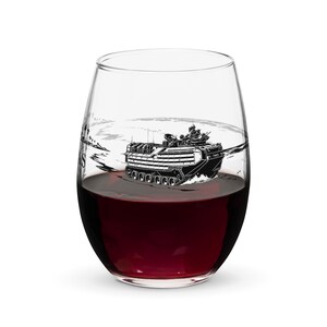 May include: A clear stemless wine glass filled with deep red wine. The glass features a black and white graphic illustration of an amphibious assault vehicle moving through water, with partial text visible on the side of the glass.