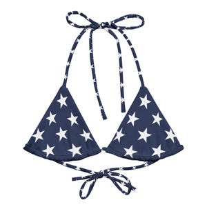 May include: A navy blue bikini top featuring a white star pattern throughout. The garment includes two triangular cups, thin adjustable neck ties, and a matching tie closure at the center back, all decorated with the same white star print design.