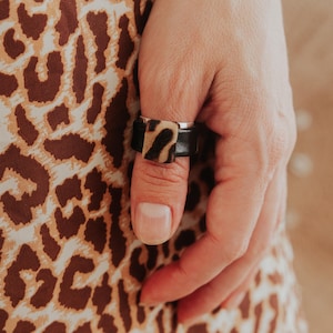 May include: A black leather ring with a silver band and a square leopard print accent. The ring is worn on a finger, with a leopard print fabric in the background.