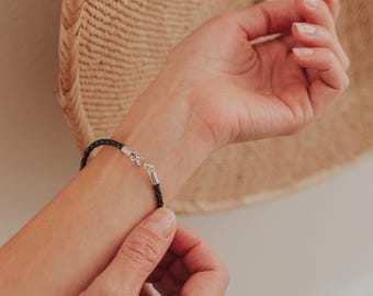 以下が含まれることがあります： 黒い編み込みレザーブレスレット、シルバーの留め金付き。 ブレスレットは手首に留められています。 背景には、織られたバスケットが含まれています。