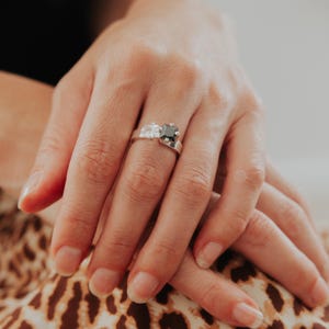 May include: A silver ring with two square gemstones, a white and a black one. The ring has a contemporary design, with the stones placed next to each other. The ring is displayed on a hand, highlighting the jewellery's design and style.
