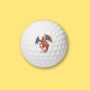 May include: White golf ball featuring a cartoon Charizard design in orange, red, and blue. The golf ball has a dimpled surface and the text "No. 0006" is printed below the graphic. The background is a solid yellow.