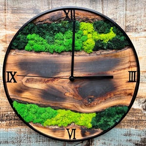 May include: A round wooden clock with Roman numeral markings and a black metal frame. The clock face is decorated with green moss and a dark wood grain pattern.