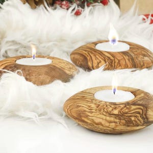 Könnte beinhalten: Drei hölzerne Kerzenhalter mit brennenden weißen Kerzen. Die Halter sind oval geformt und haben ein natürliches Holzmaserungsmuster. Sie liegen auf einem Bett aus weißem Kunstfell, mit Andeutungen von Rot und Braun im Hintergrund.