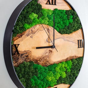 May include: A round wall clock with a black frame and a natural wood grain face. The clock features preserved green moss in the upper and lower sections, with black Roman numerals at the XII, III, VI, and IX positions and black hands.