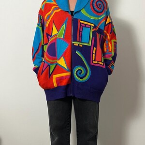 May include: A colorful zip-up sweater with a vibrant geometric design. The sweater features a mix of red, blue, yellow, and purple, with a large collar and ribbed cuffs. The design includes abstract shapes and patterns.