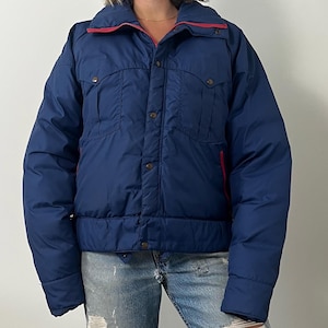 May include: A navy blue puffer jacket with red trim on the collar and zipper. The jacket has two chest pockets and a button-up front. The jacket is worn with blue jeans.