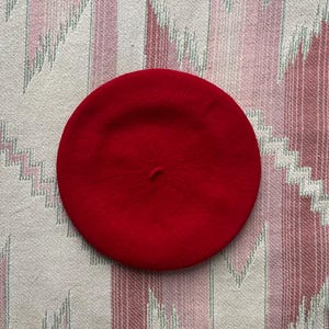 May include: A bright red beret, a classic round hat made of felt. The beret is a solid, vibrant color and sits on a patterned fabric background with pink, white, and green hues.