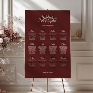 Puede incluir: Un cartel rectangular de color rojo oscuro para la distribución de mesas de boda sobre un caballete de metal dorado. El cartel presenta texto en blanco que dice A PLACE For You, PLEASE FIND YOUR SEAT, y enumera las mesas del uno al doce con nombres, terminando con SOPHIA & NATHAN.