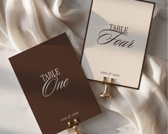 May include: Two rectangular table number cards held by gold metal stands. One card is dark brown with white text reading TABLE One and SOPHIA & NATHAN. The other is cream with dark brown text reading TABLE Four and SOPHIA & NATHAN.