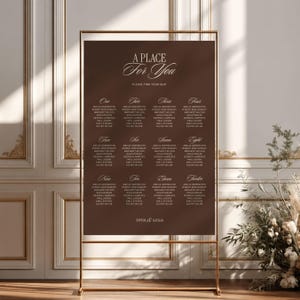 May include: A dark brown rectangular wedding seating chart sign displayed on a thin gold metal frame stand. The sign features the text A PLACE FOR YOU, PLEASE FIND YOUR SEAT, and lists twelve tables with names in white serif and script fonts.