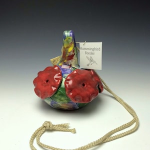 May include: A colorful ceramic hummingbird feeder with red flower-shaped feeding ports and a hanging cord. The feeder has a tag that reads "Hummingbird Feeder".