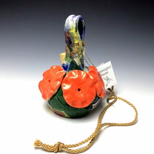 May include: A handmade ceramic hummingbird feeder with a colorful floral design and three red flower-shaped openings. It has a brown cord for hanging.