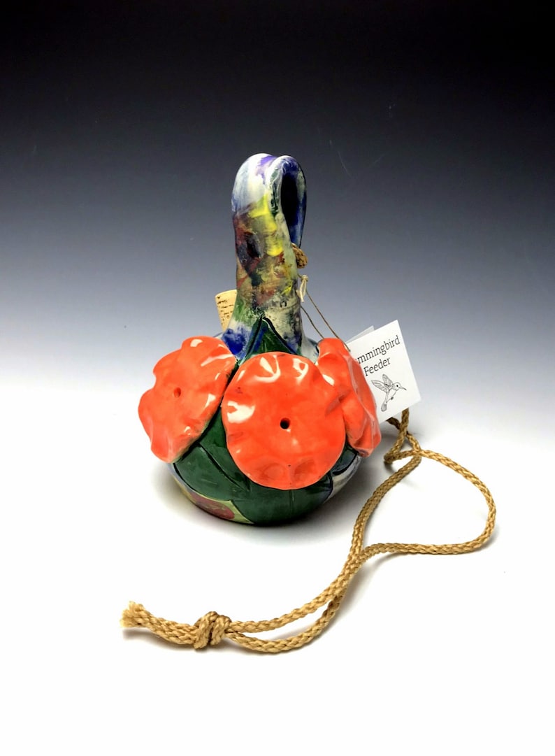 May include: A handmade ceramic hummingbird feeder with a colorful floral design and three red flower-shaped openings. It has a brown cord for hanging.