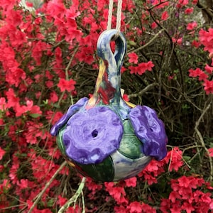 May include: A colorful ceramic hanging ornament with purple flowers and green leaves. The ornament is painted with a variety of colors, including blue, yellow, and red.