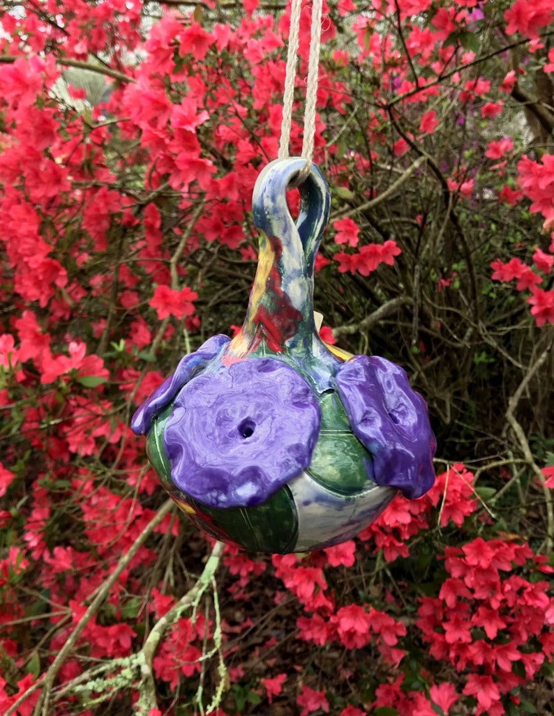 May include: A colorful ceramic hanging ornament with purple flowers and green leaves. The ornament is painted with a variety of colors, including blue, yellow, and red.