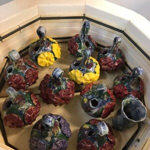 May include: A collection of 12 ceramic ornaments in a kiln. The ornaments are hand-painted with various floral and geometric patterns in shades of red, blue, yellow, and green.