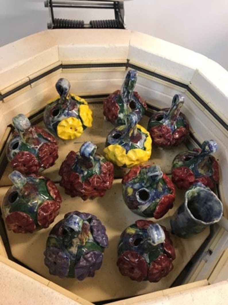 May include: A collection of 12 ceramic ornaments in a kiln. The ornaments are hand-painted with various floral and geometric patterns in shades of red, blue, yellow, and green.