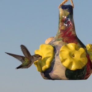 May include: A hummingbird hovers near a colorful ceramic hummingbird feeder with yellow flower-shaped perches. The feeder is decorated with blue, green, red, and white paint.