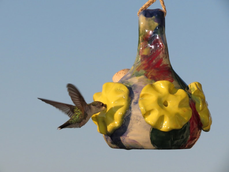 May include: A hummingbird hovers near a colorful ceramic hummingbird feeder with yellow flower-shaped perches. The feeder is decorated with blue, green, red, and white paint.