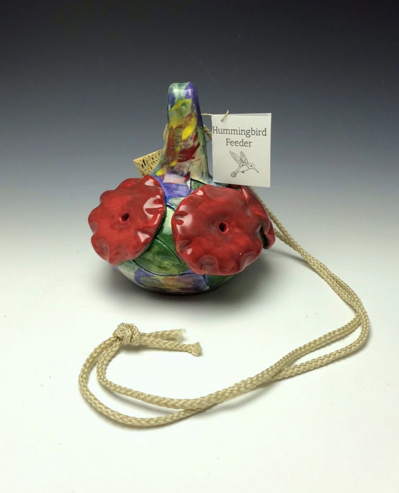 May include: A colorful ceramic hummingbird feeder with red flower-shaped feeding ports and a hanging cord. The feeder has a tag that reads "Hummingbird Feeder".