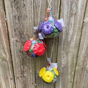 May include: Three ceramic bird feeders hanging from a wooden fence. The feeders are decorated with colorful flowers and have a hole in the bottom for birds to access the food. The feeders are purple, red, and yellow.