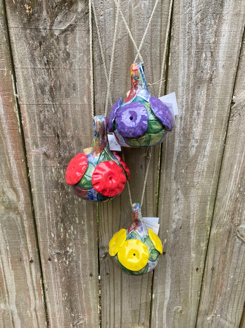 May include: Three ceramic bird feeders hanging from a wooden fence. The feeders are decorated with colorful flowers and have a hole in the bottom for birds to access the food. The feeders are purple, red, and yellow.