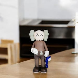 May include: A brown and tan vinyl figure with a skull-like face and X-shaped eyes. The figure has white gloves and holds a blue accessory. The figure is standing on a wooden surface.