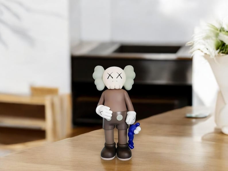May include: A brown and tan vinyl figure with a skull-like face and X-shaped eyes. The figure has white gloves and holds a blue accessory. The figure is standing on a wooden surface.