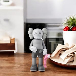 May include: A gray vinyl figure with a skull-like head and crossed-out eyes. The figure is wearing gray shorts, gloves, and shoes, and is holding a pink object. The figure is standing on a wooden surface.