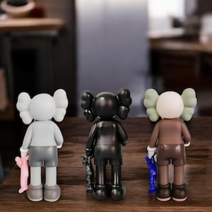 May include: Three collectible vinyl figures in a row, each with a unique color scheme. The figures have a distinctive design with large, rounded ears and hold a small companion. The figures are standing on a wooden surface.