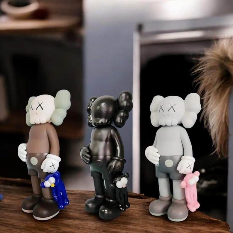 May include: Three KAWS Companion figures in brown, black, and gray. Each figure has a distinctive head with X-shaped eyes and a rounded body. The figures are holding small plush companions in blue and pink.