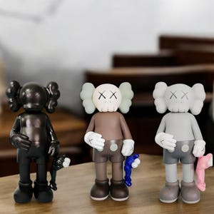 May include: Three collectible vinyl figures in black, brown, and gray. Each figure has a unique design with X-shaped eyes and a rounded head. The figures are holding accessories, including a black, blue, and pink item.