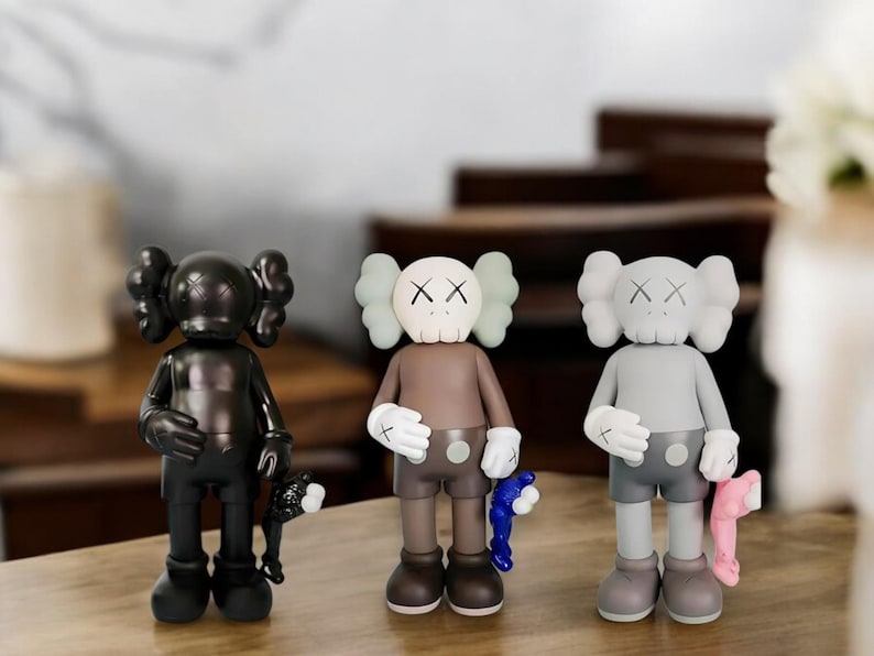 May include: Three collectible vinyl figures in black, brown, and gray. Each figure has a unique design with X-shaped eyes and a rounded head. The figures are holding accessories, including a black, blue, and pink item.