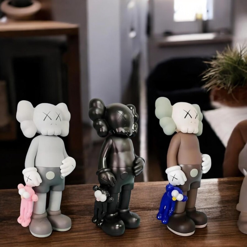 May include: Three KAWS Companion figures in a row. The figures are in different colors: gray, black, and brown. Each figure is holding a small plush toy. The figures have a distinctive skull-like face with X-shaped eyes and a round head.