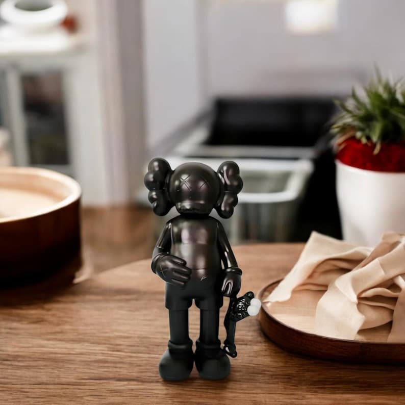 May include: A black vinyl figurine of a cartoon character with large, rounded ears and crossed-out eyes. The figure is holding a smaller, matching character. The figurine is standing on a wooden surface.