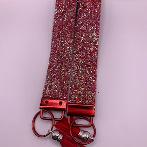 May include: Two red glitter wrist keychains with red metal hardware and a red tassel. The keychains have a rectangular shape and are covered in sparkling glitter. The keychains are connected to a red metal ring.