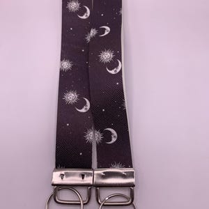 May include: Two black keychains with a celestial pattern of white suns, moons, and stars. The keychains have silver metal hardware and large silver rings for keys.