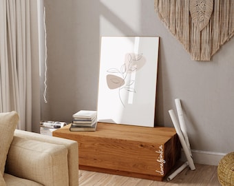 May include: A minimalist line art print of a branch with leaves and abstract shapes in shades of pink and brown. The print is framed and hanging on a wall above a wooden shelf.