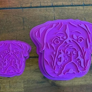 May include: Two purple plastic cookie cutters in the shape of a dog's face. The larger cutter is approximately 4 inches wide, and the smaller one is about 2 inches wide. The dog's face is detailed with eyes, nose, and wrinkles.