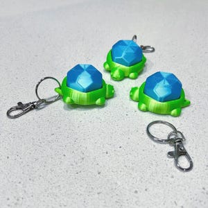 May include: Three 3D-printed turtle keychains. Each keychain features a green turtle body and a blue, geometric shell. The keychains have a silver keyring and a lobster clasp for easy attachment to bags or keys.