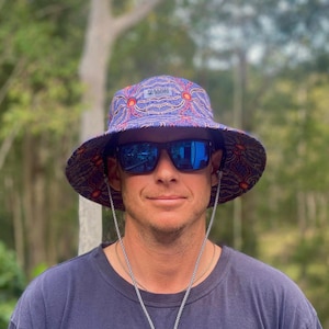 Könnte beinhalten: Eine Person trägt einen blauen und roten gemusterten Bucket Hat mit schwarzem Rand und schwarzem Kinnriemen. Der Hut hat den Text "MAD ABOUT" auf der Vorderseite aufgedruckt. Die Person trägt auch eine blaue Sonnenbrille.