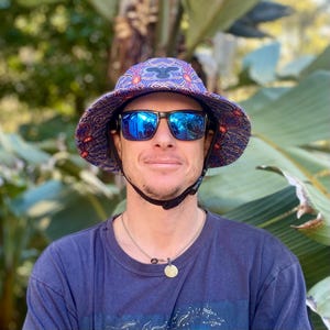 May include: A person wearing a blue and red patterned bucket hat with a black chin strap and blue mirrored sunglasses. The hat has a black and white design on the front.