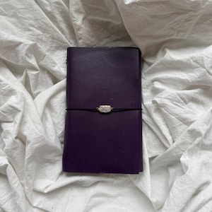 May include: A rectangular dark purple leather journal secured with a thin black elastic cord. The cord features a small, iridescent, rectangular-shaped pearl bead centered horizontally across the front cover of the notebook.