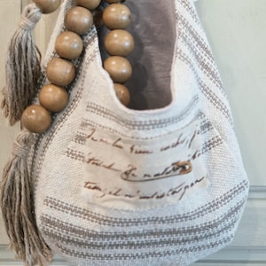 May include: A hanging fabric storage pouch in off-white with beige stripes, featuring a front pocket with cursive script text, a decorative wooden bead strand with a jute tassel, and a light gray interior lining, suspended by a fabric loop.