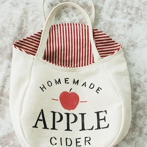 May include: Cream-colored canvas tote bag with a red and white striped interior. The bag features a red apple graphic and the words "Homemade Apple Cider" in black lettering. It has a small handle.